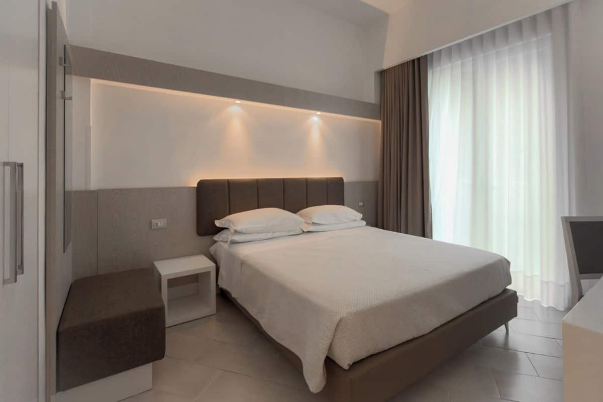 Una habitación de hotel moderna con una cama grande, mesitas de noche, televisor integrado y un amplio ventanal con cortinas.
