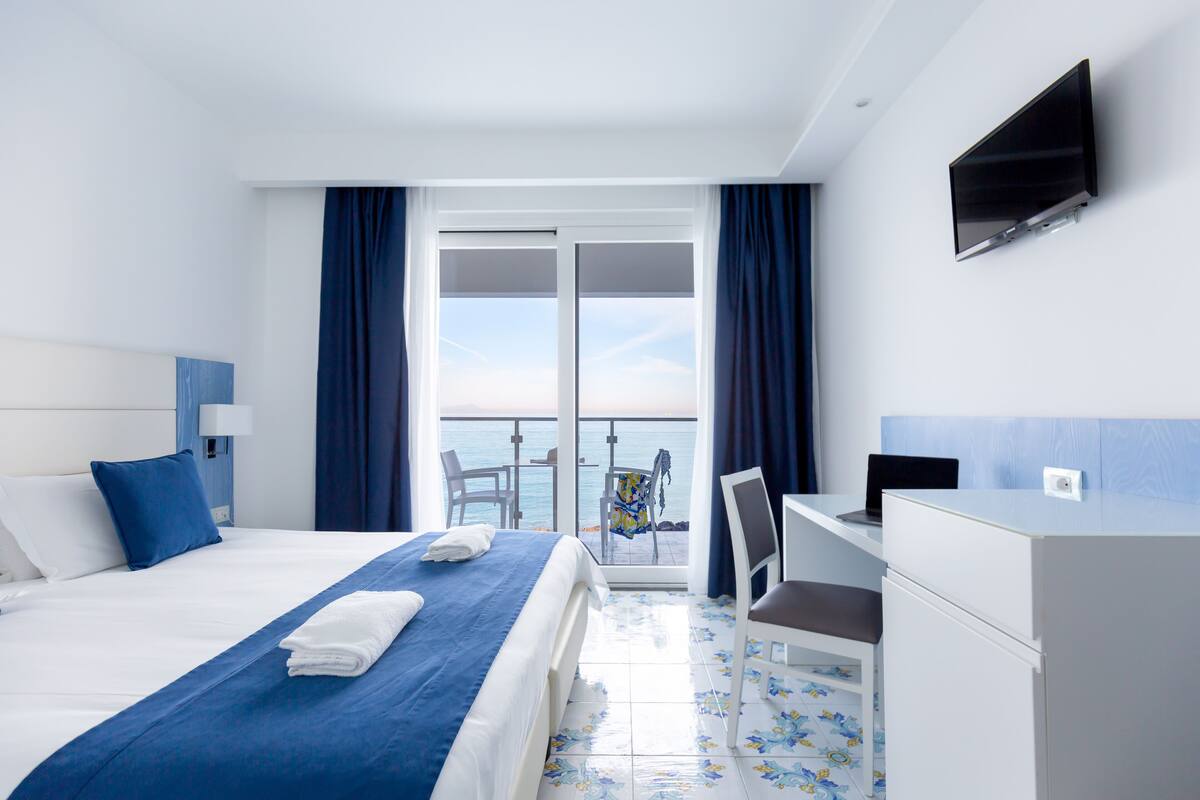 Habitación de hotel con cama, escritorio, silla, televisión y balcón con vista al mar.