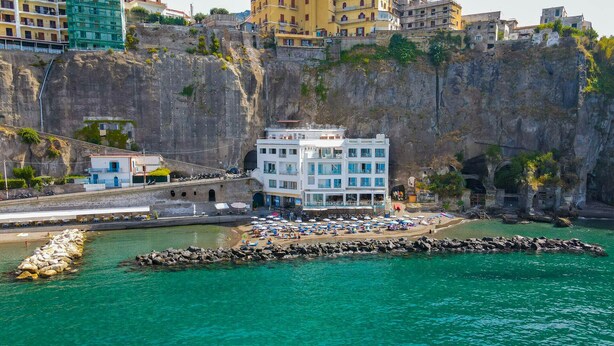 Un hotel costero con un edificio grande, una costa rocosa y un mar azul cristalino.