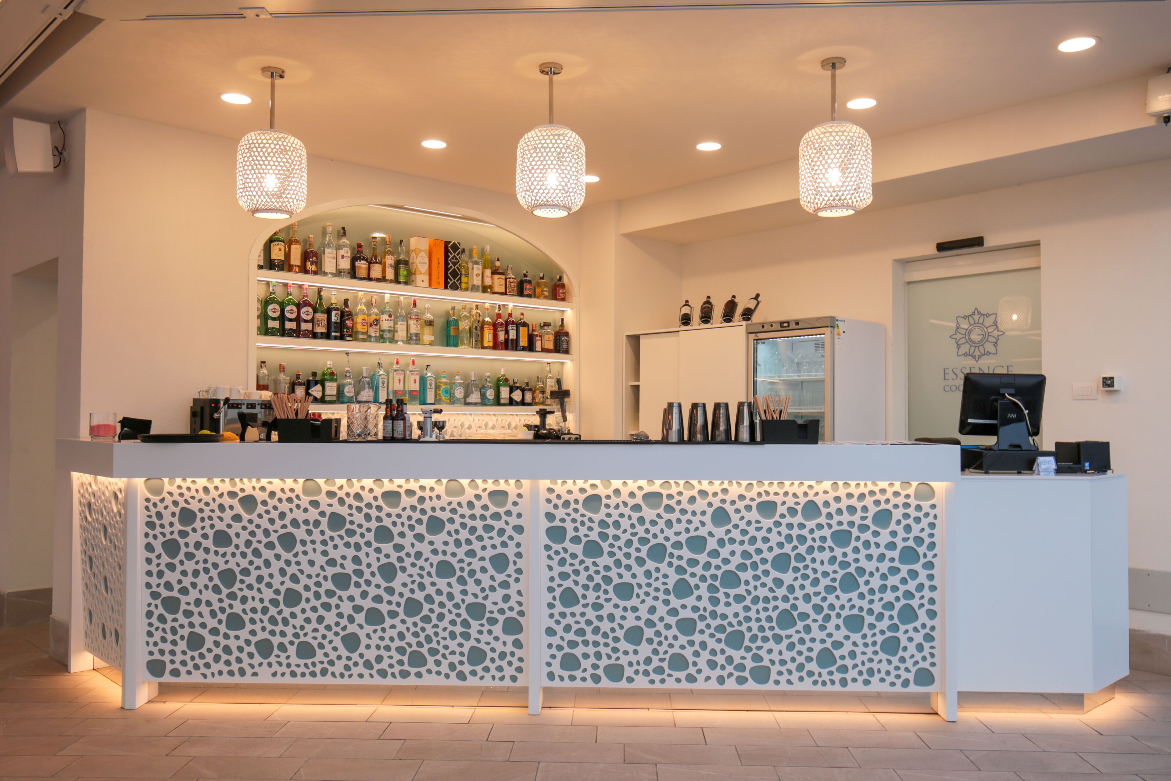 Zona de bar con mostrador blanco, estantes con botellas y tres luces colgantes.