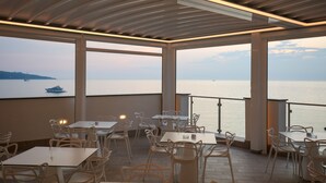 Un restaurante con mesas y sillas, ventanas amplias y vista al mar.