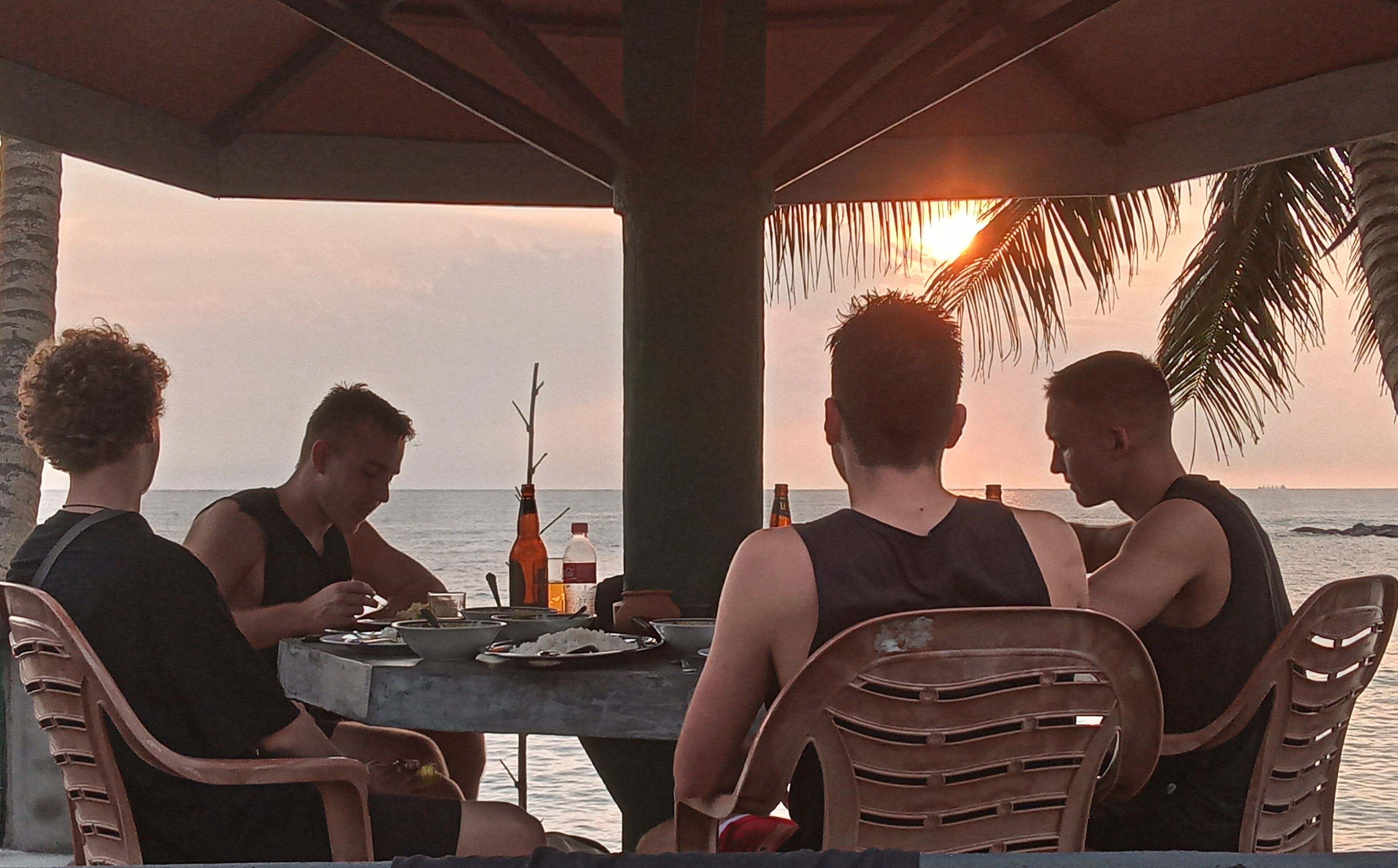 A group of people dining at an outdoor table with a view of the sunset over the ocean.