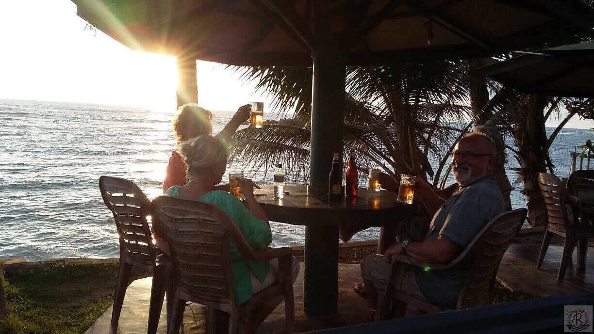 Un groupe de personnes est assis à une table en plein air, d’où l’on peut admirer l’océan au coucher du soleil.