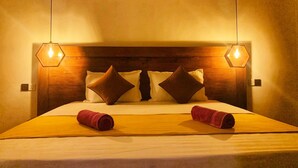 Habitación de hotel con cabecera de madera, dos lámparas colgantes hexagonales, una cama con ropa blanca y dos toallas enrolladas sobre la cama.