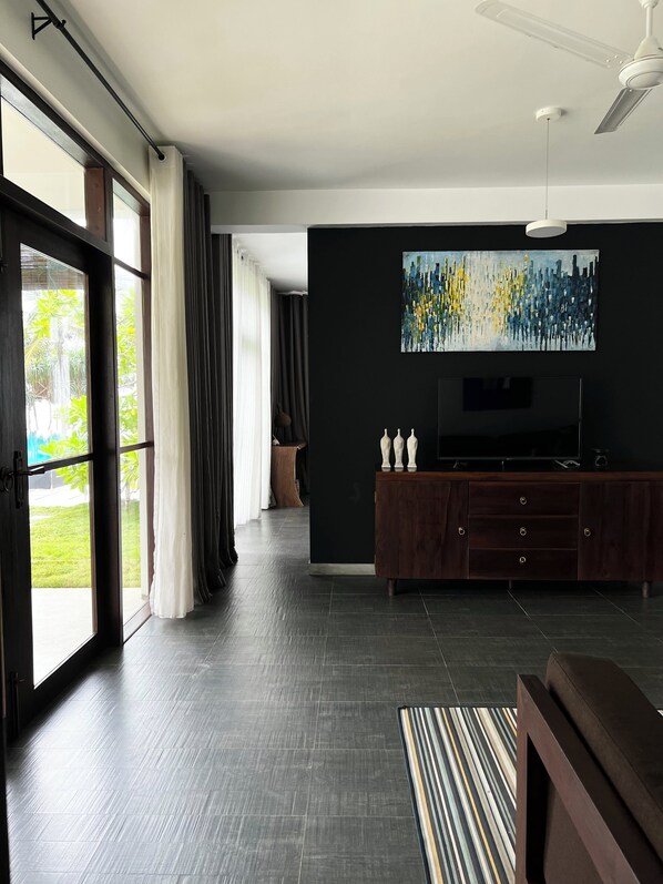 A modern living room with a dark floor, a wooden cabinet, a flat-screen TV, and a ceiling fan. - Mandala Villas (Rekawa)