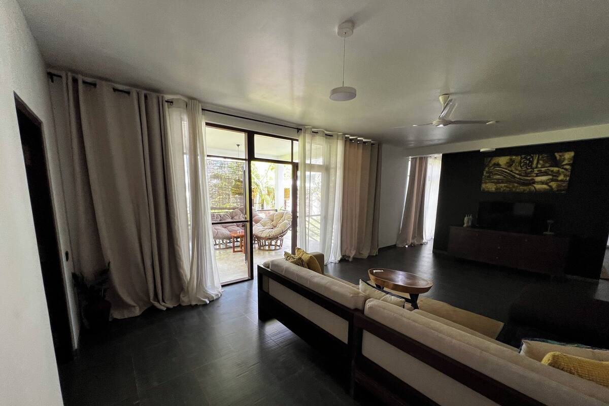 A modern living room with a sofa, a coffee table, and a TV. There is a sliding glass door leading to an outdoor area with a chair and a table.