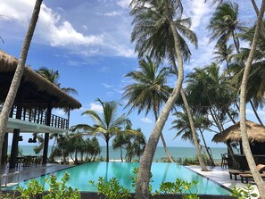 A poolside area with a clear view of the ocean, surrounded by palm trees and thatched-roof structures. - Mandala Villas (Rekawa)