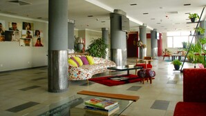 Un hall d’entrée spacieux, avec un canapé, une table basse et un fauteuil rouge. On y trouve de grandes colonnes, un tapis rouge et une bibliothèque remplie de livres.