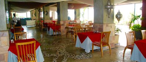 Un amplio comedor interior con varias mesas cubiertas con manteles rojos y sillas de madera. El suelo presenta un original diseño de mosaico.
