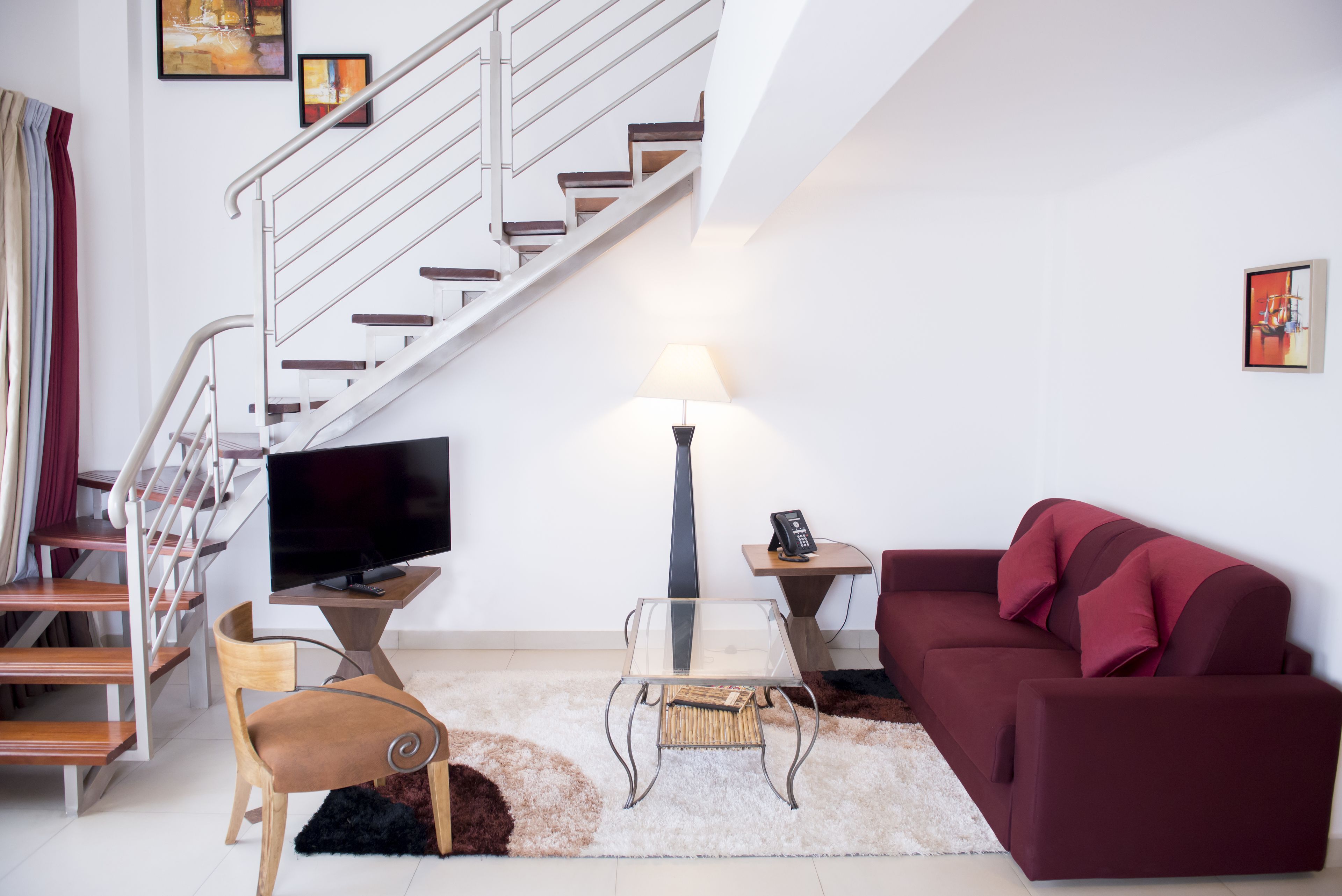 A living room with a staircase, a flat-screen TV, a glass coffee table, a red sofa, and a lamp.