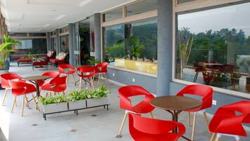 Um espaço amplo e interno, com cadeiras e mesas vermelhas, grandes janelas com vista para o verde, e um design arquitetônico moderno.