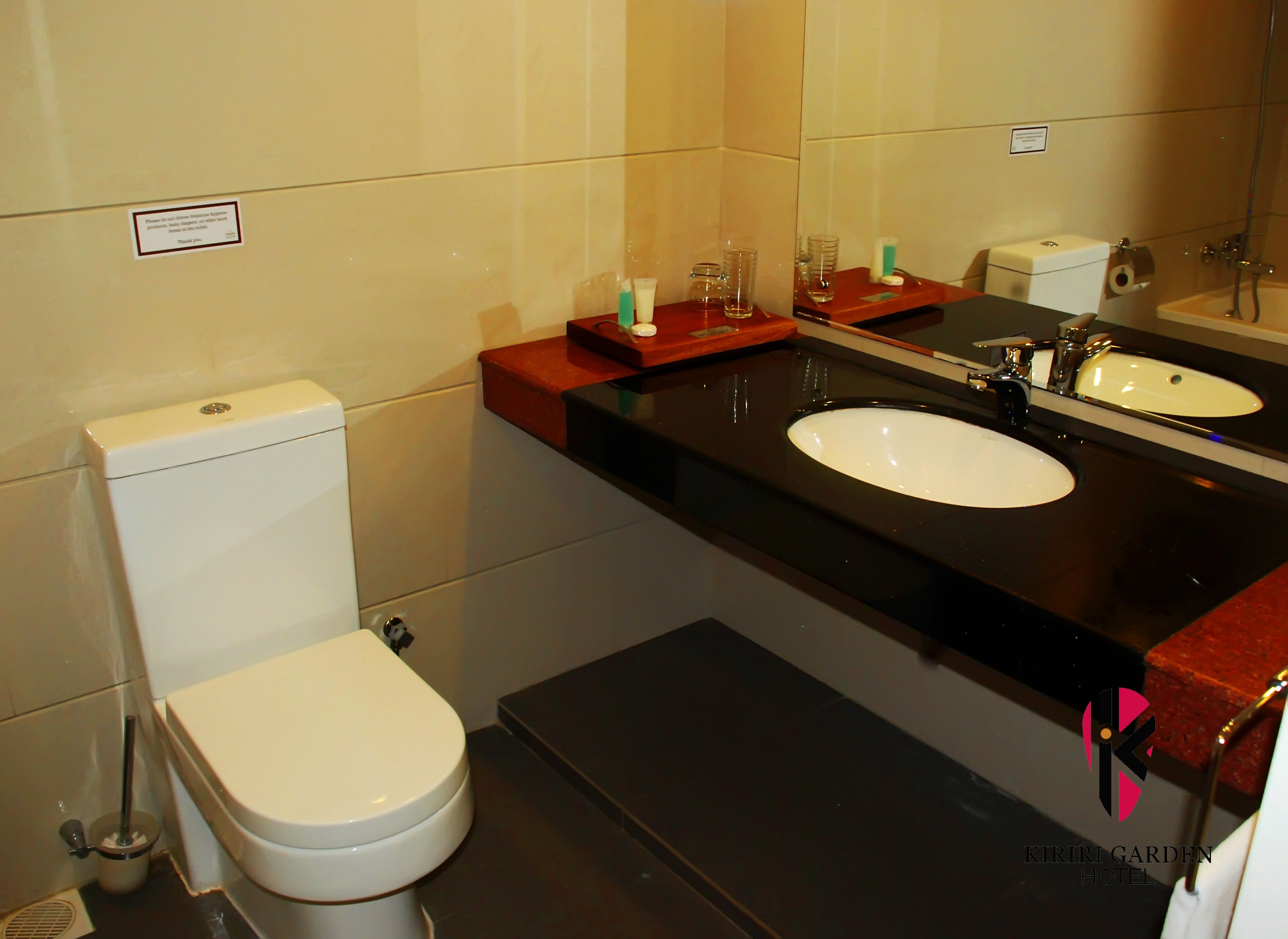 A bathroom with a toilet, a sink, and a mirror. There is a sign on the wall and a small shelf with toiletries.