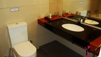A bathroom with a toilet, a sink, and a mirror. There is a sign on the wall and a small shelf with toiletries.