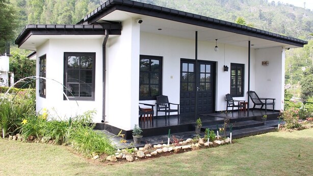 A modern white building with a black roof, a covered porch with chairs, and a stone pathway leading to the entrance. - Royal Eco Resort (Haldummulla)