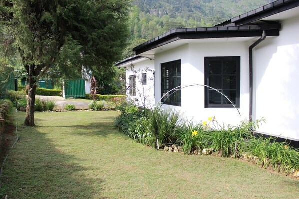 A white building with black-framed windows, surrounded by a well-maintained garden with various plants and a tree. - Royal Eco Resort (Haldummulla)