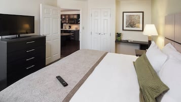 A hotel room with a bed, a dresser, a television, a desk with a lamp, and a kitchenette in the background.