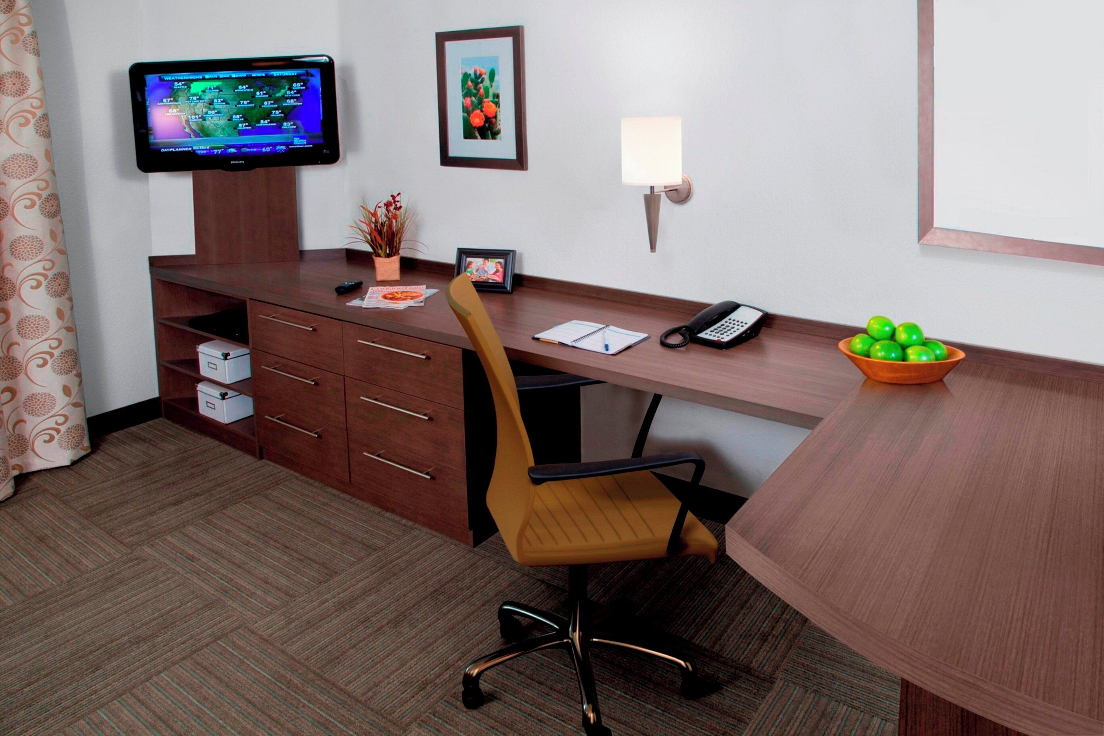 Habitación de hotel con un escritorio de madera, una silla, un televisor, un cuadro enmarcado, una lámpara, un teléfono, un frutero y una pequeña planta.