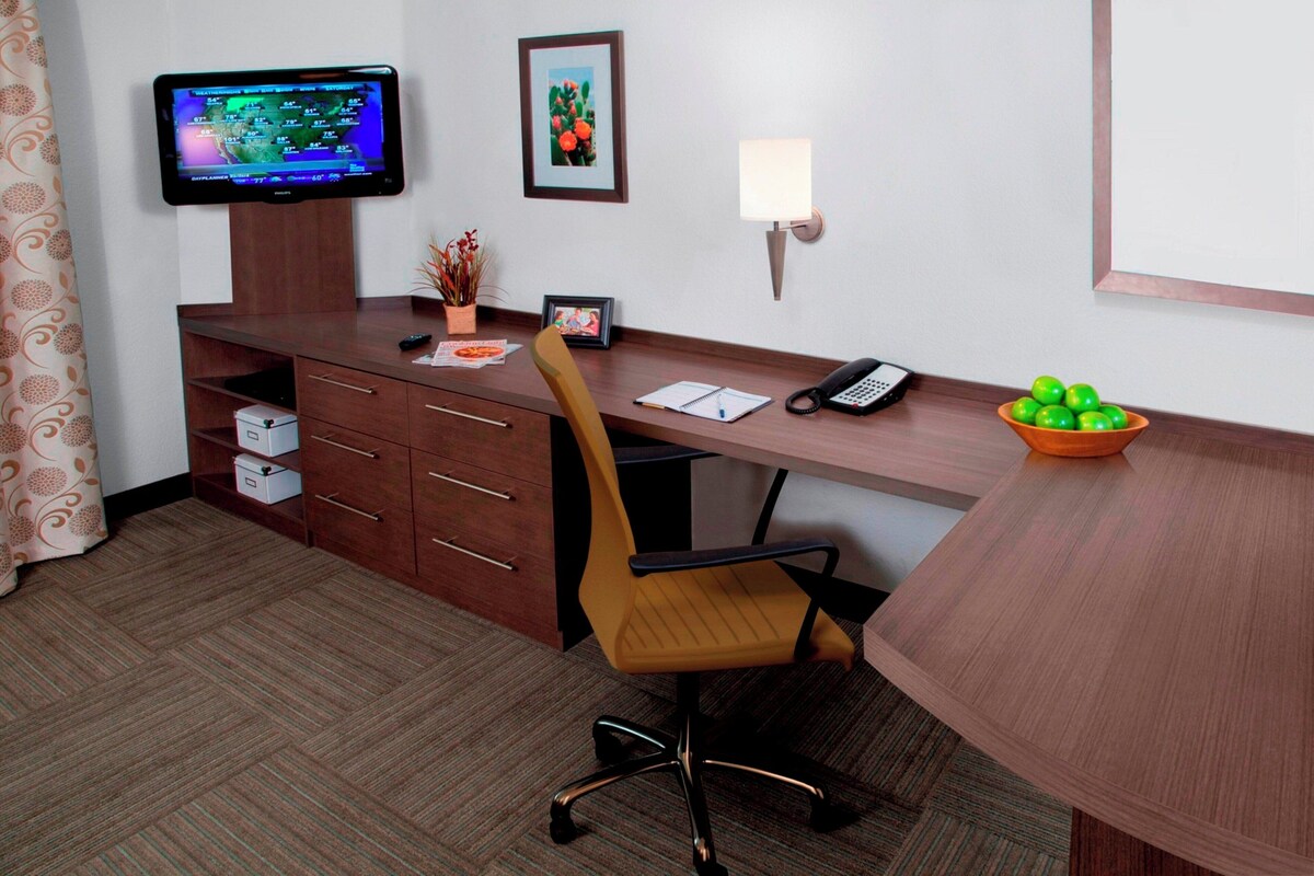 A hotel room with a wooden desk, a chair, a television, a framed picture, a lamp, a telephone, a bowl of fruit, and a small plant.
