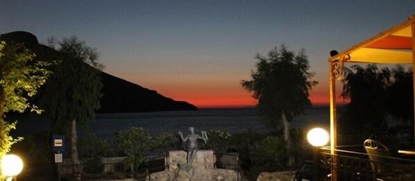 Vista del atardecer desde una terraza, con una estatua en primer plano, un farol y un edificio al fondo.