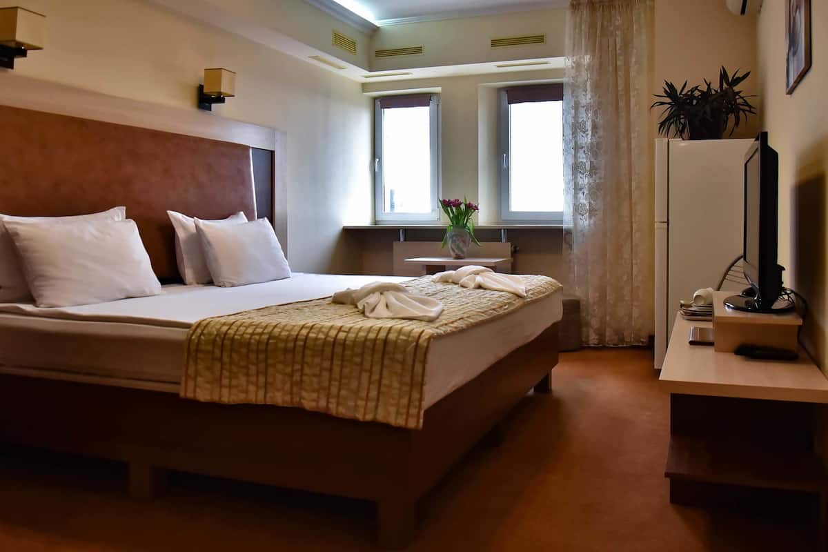 A hotel room with a large bed, a bedside table with a vase of flowers, a desk with a computer, and a window with curtains.
