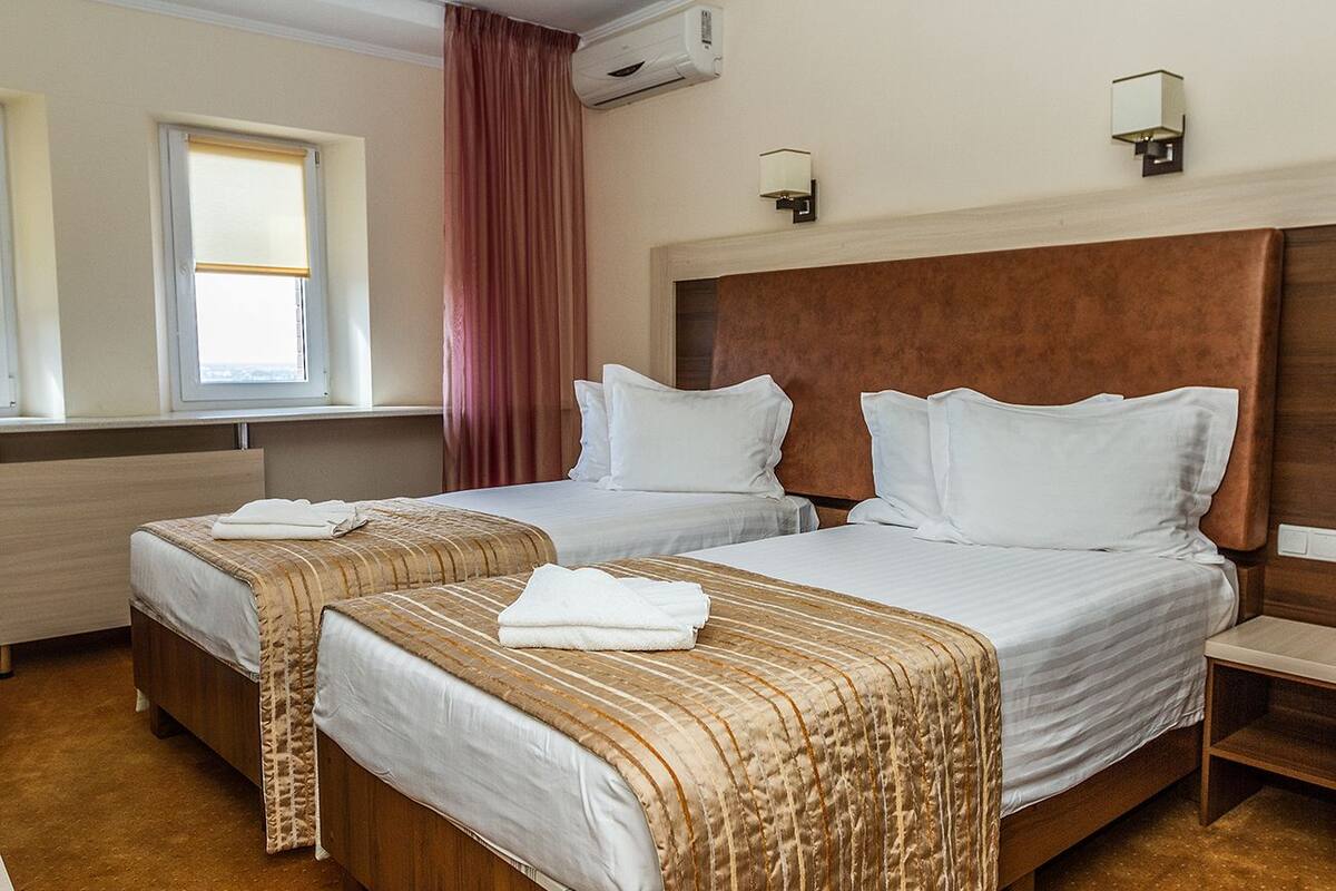 A hotel room with two beds, a window with blinds, an air conditioning unit, and a nightstand.