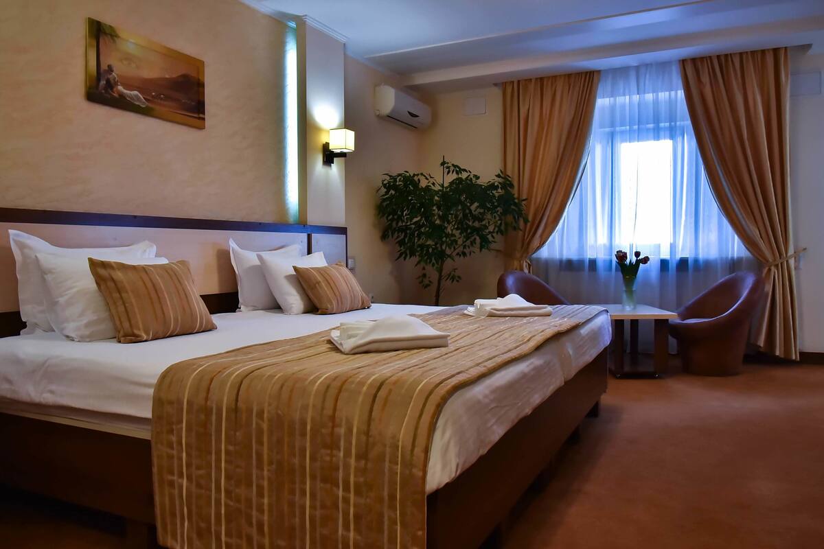 A hotel room with a large bed, a small table, a chair, a potted plant, and a painting on the wall.