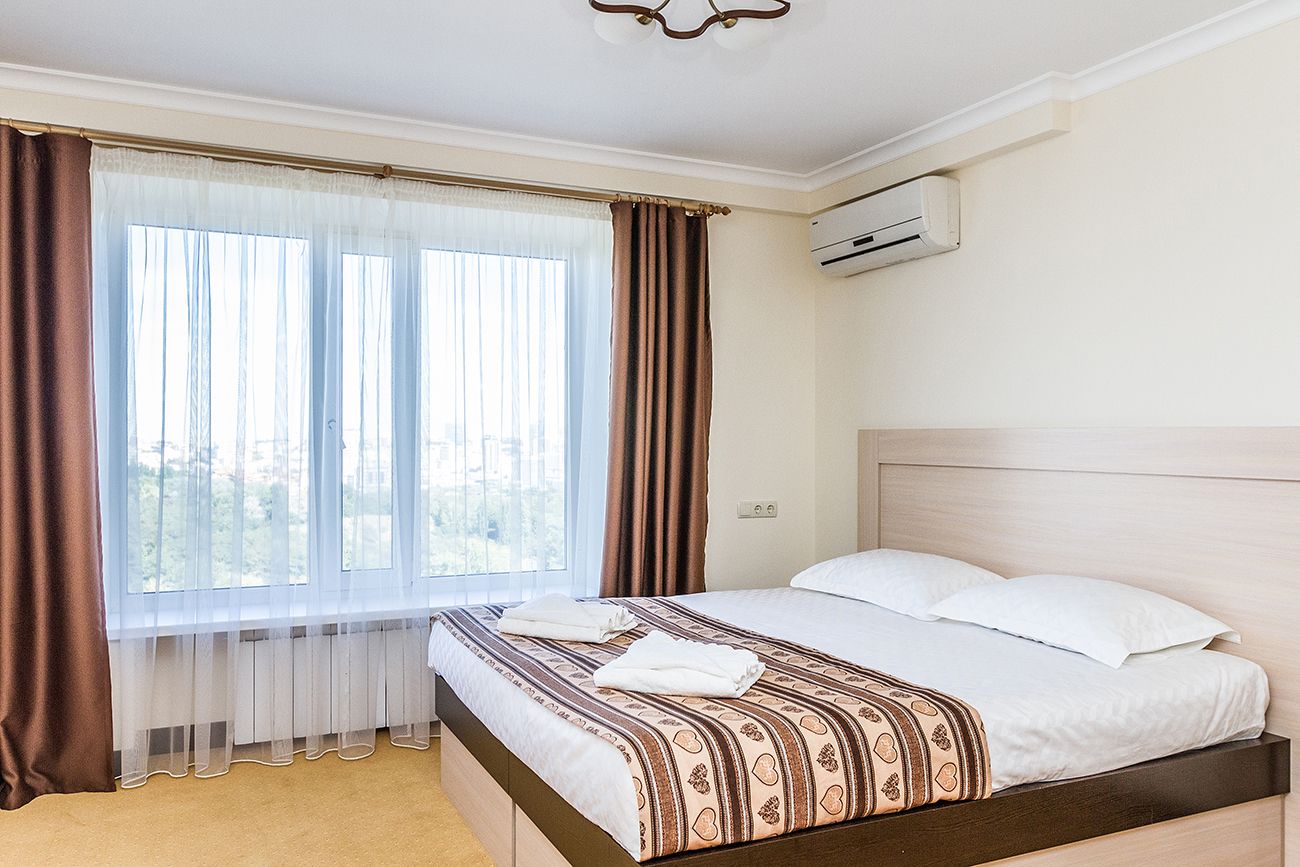 A hotel room with a bed, a window with curtains, an air conditioning unit, and a view of the outdoors.