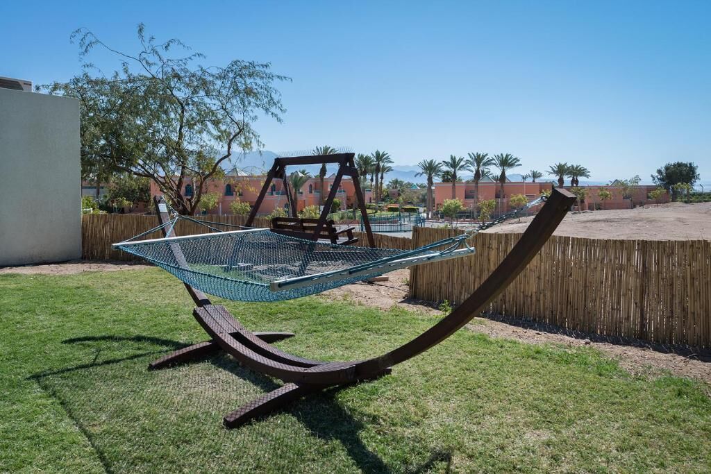 A hammock chair with a netted seat, set on a grassy lawn with a wooden fence and palm trees in the background.