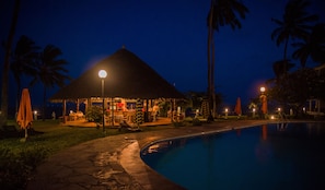 A nighttime scene of a resort with a pool, a thatched-roof bar area, and palm trees. - Nyali Beach Holiday Resort (Mombasa)