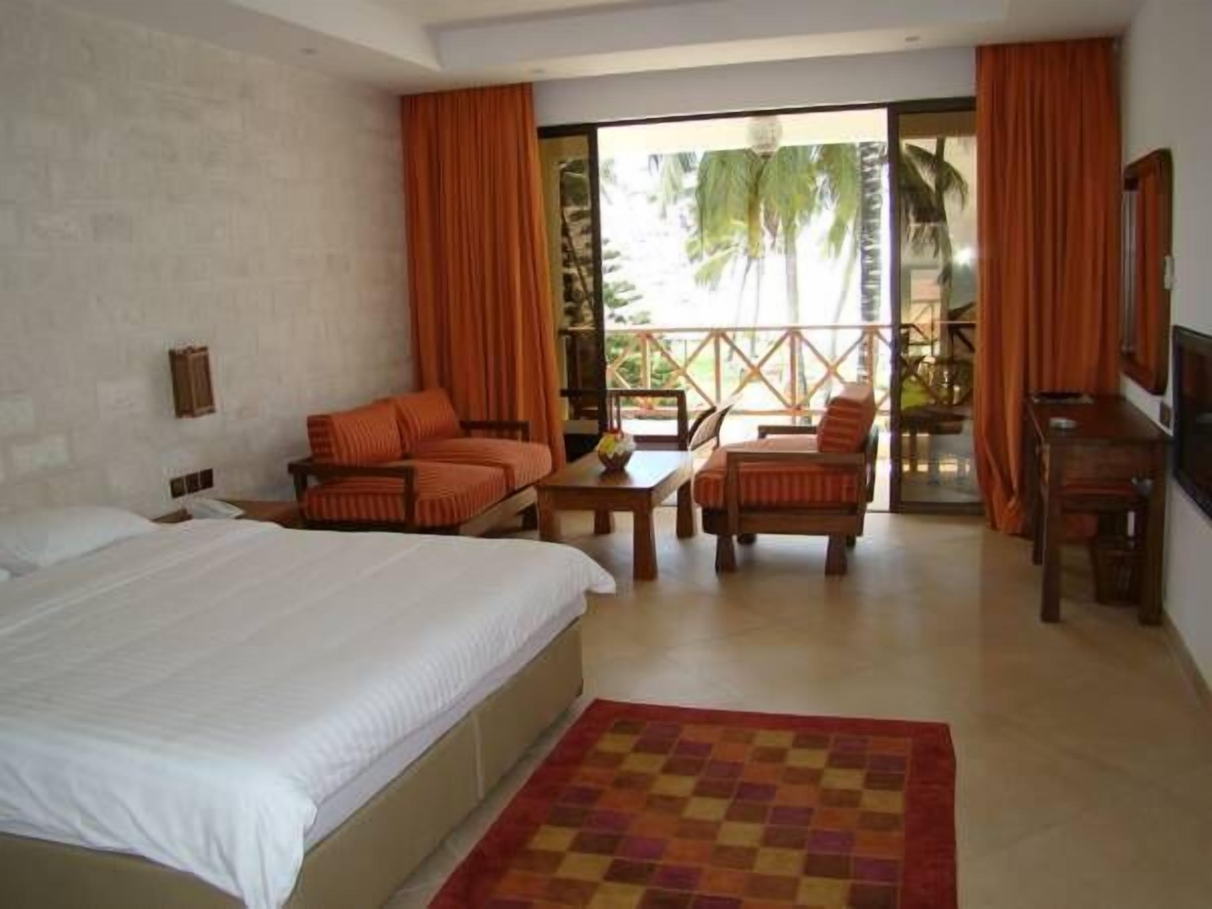 Une chambre d’hôtel avec un lit, un canapé, une table et une chaise. Elle dispose d’un balcon avec vue sur les palmiers.