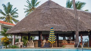 A thatched-roof structure with a pool in front, surrounded by palm trees and chairs. - Nyali Beach Holiday Resort (Mombasa)