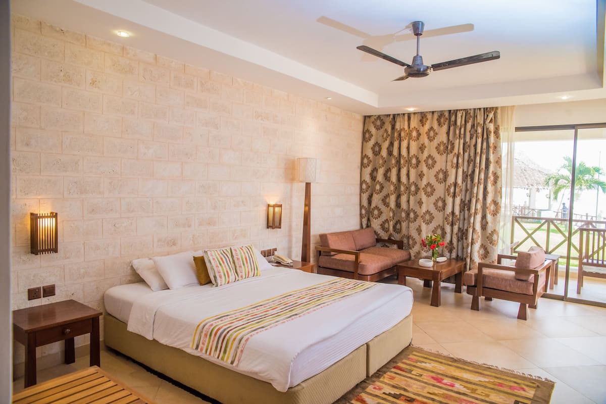 A hotel room with a brick wall, a bed with white bedding, a ceiling fan, a balcony with a view, and a patterned curtain.