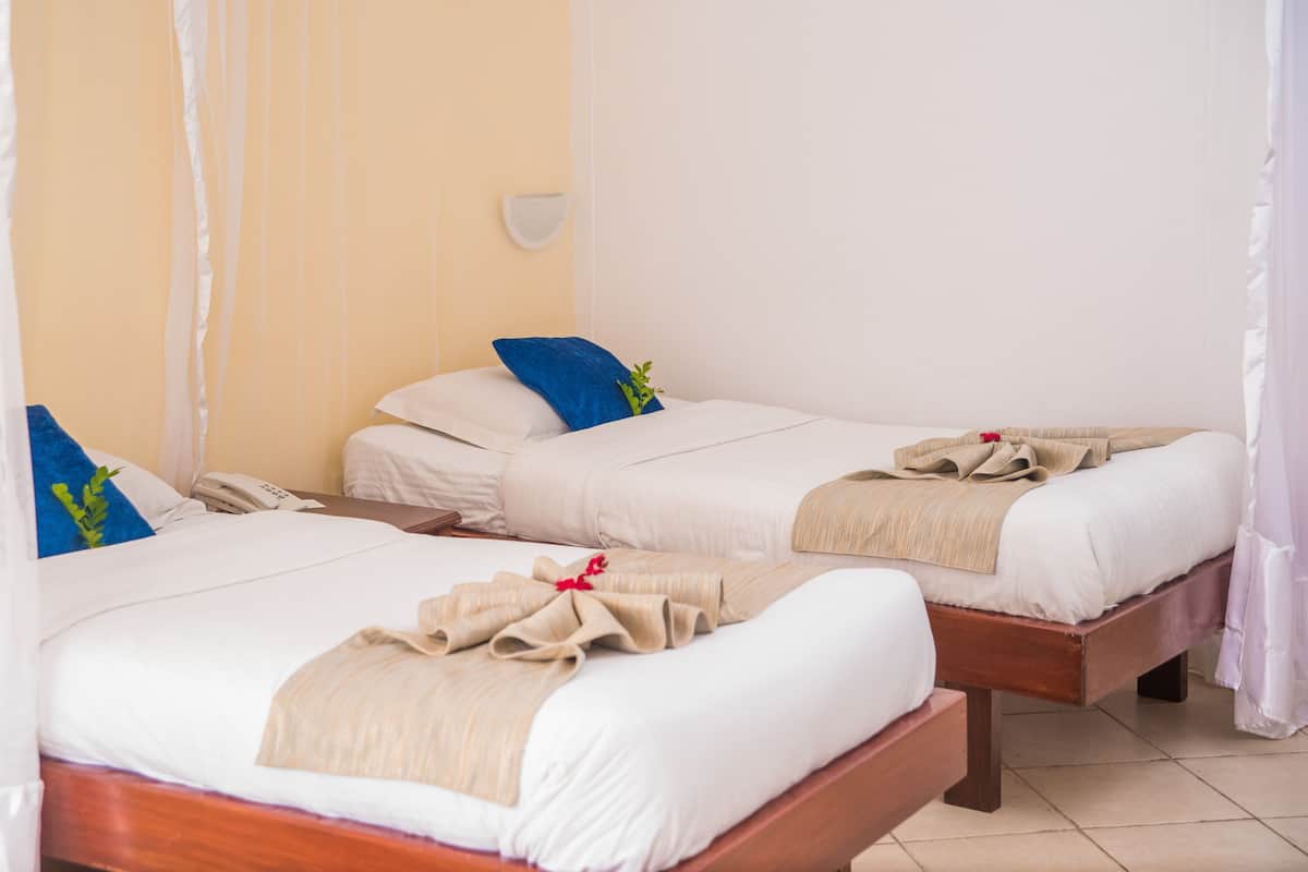 A hotel room with two beds, each with a white bedspread, a blue pillow, and a beige blanket. There is a bedside table with a phone and a brown wallet.