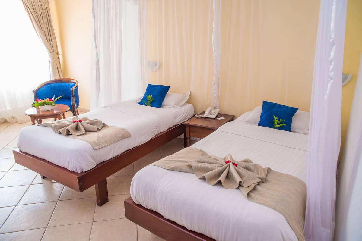 A hotel room with two beds, each with a wooden frame and white bedding. There is a small table with a plant and a chair in the corner.