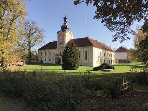 A large building with a prominent tower and a red roof, surrounded by a well-maintained lawn and trees. - Zámek Lešany (Lešany)