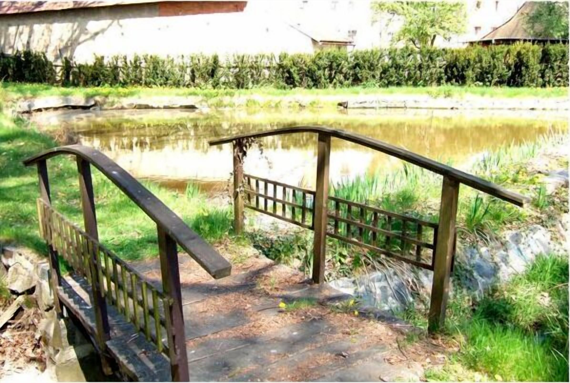 A wooden bridge over a pond with a metal railing, surrounded by greenery and a building in the background.