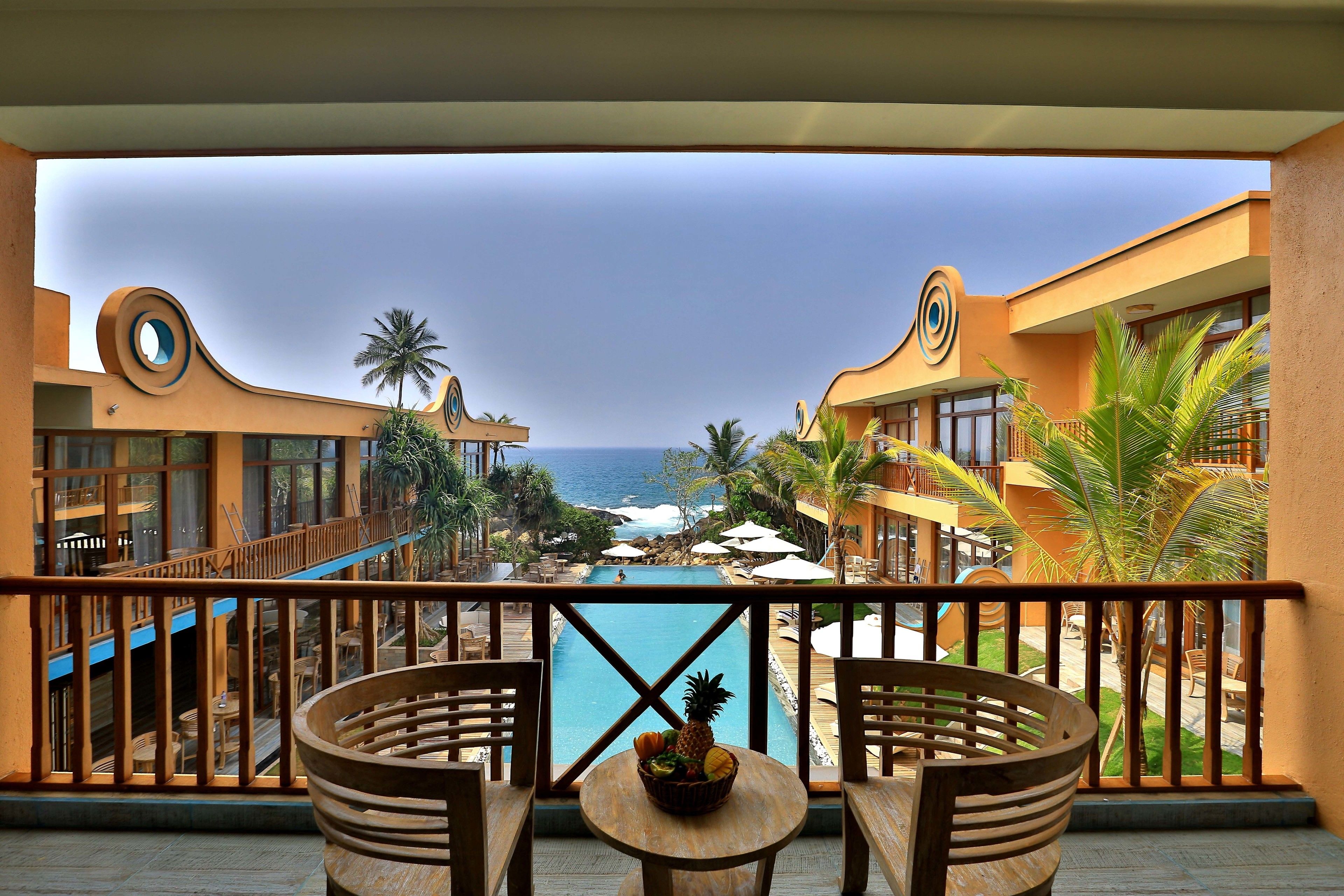 A balcony with a view of the ocean, a pool, and a tropical setting with palm trees.