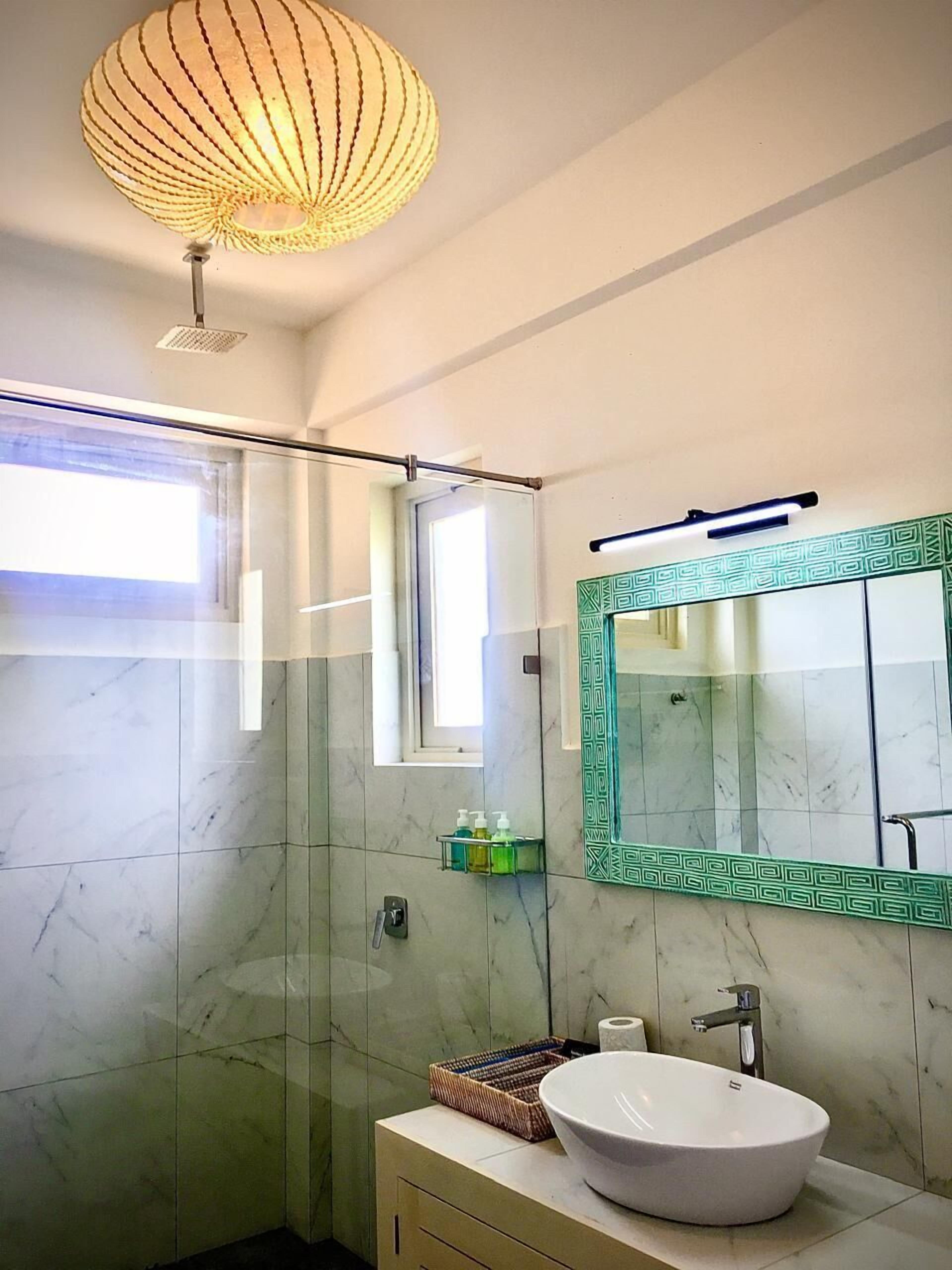 A modern bathroom with a glass shower enclosure, a white sink with a green-framed mirror, and a ceiling light fixture.