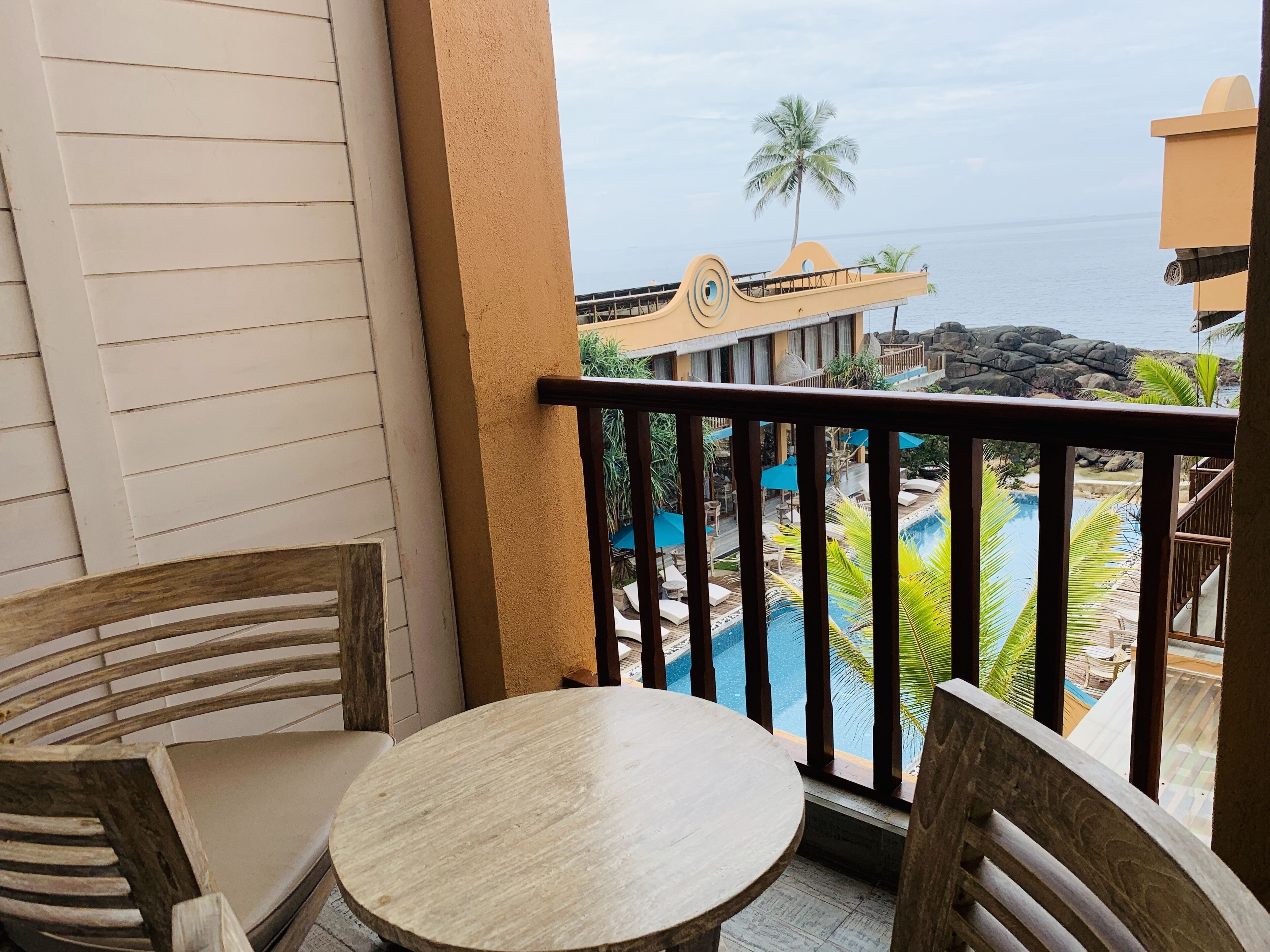 A balcony with a wooden table and chairs, overlooking a pool area with a view of the ocean and a palm tree.