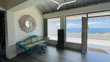 A room with a blue woven bench, a ceiling fan, and large windows offering a view of the ocean.