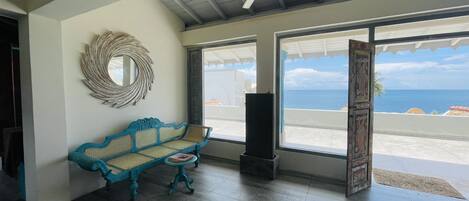 A room with a blue woven bench, a ceiling fan, and large windows offering a view of the ocean.