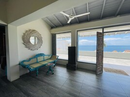 Una stanza con una panca blu intrecciata, una ventola a soffitto e ampie finestre che si affacciano sull'oceano.