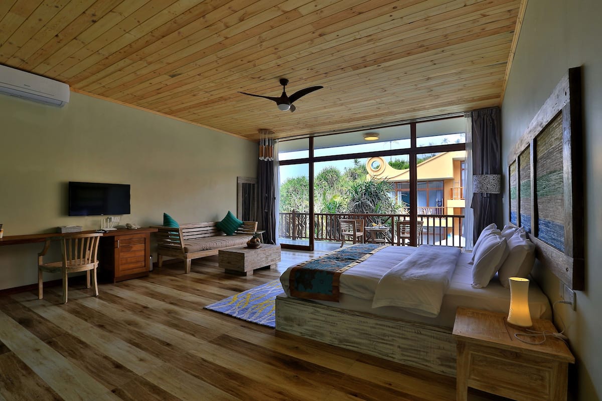 A bedroom with a wooden ceiling, a bed, a desk, a chair, and a balcony with a view of greenery.