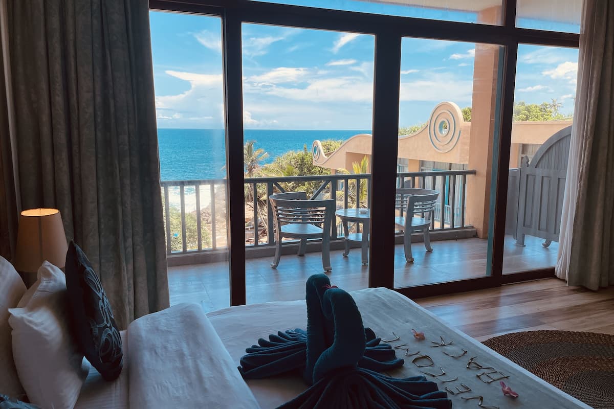 A hotel room with a large window overlooking a balcony with outdoor furniture and a view of the sea.