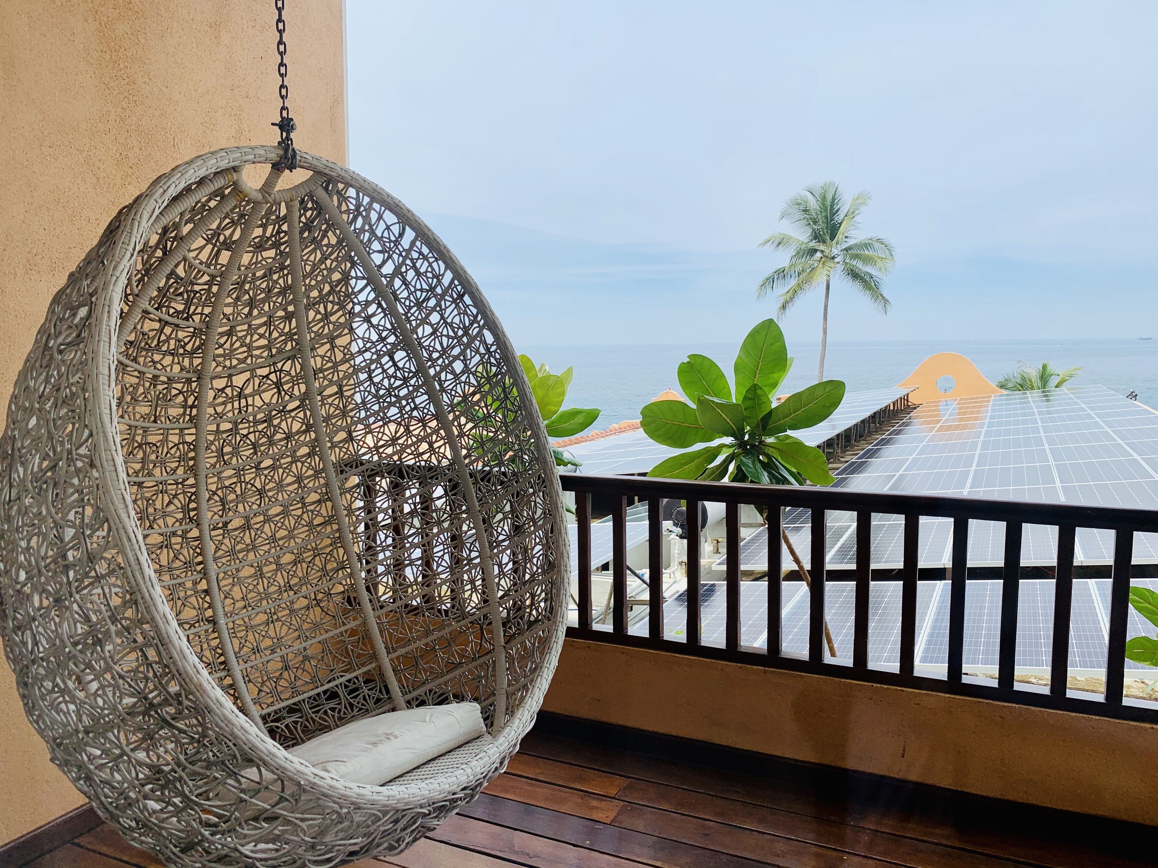 A wicker hanging chair on a balcony with a view of the sea and solar panels.