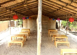 A covered outdoor seating area with wooden tables and benches, surrounded by a sandy floor and a thatched roof. - Kay's By The Sea (Vengurla)