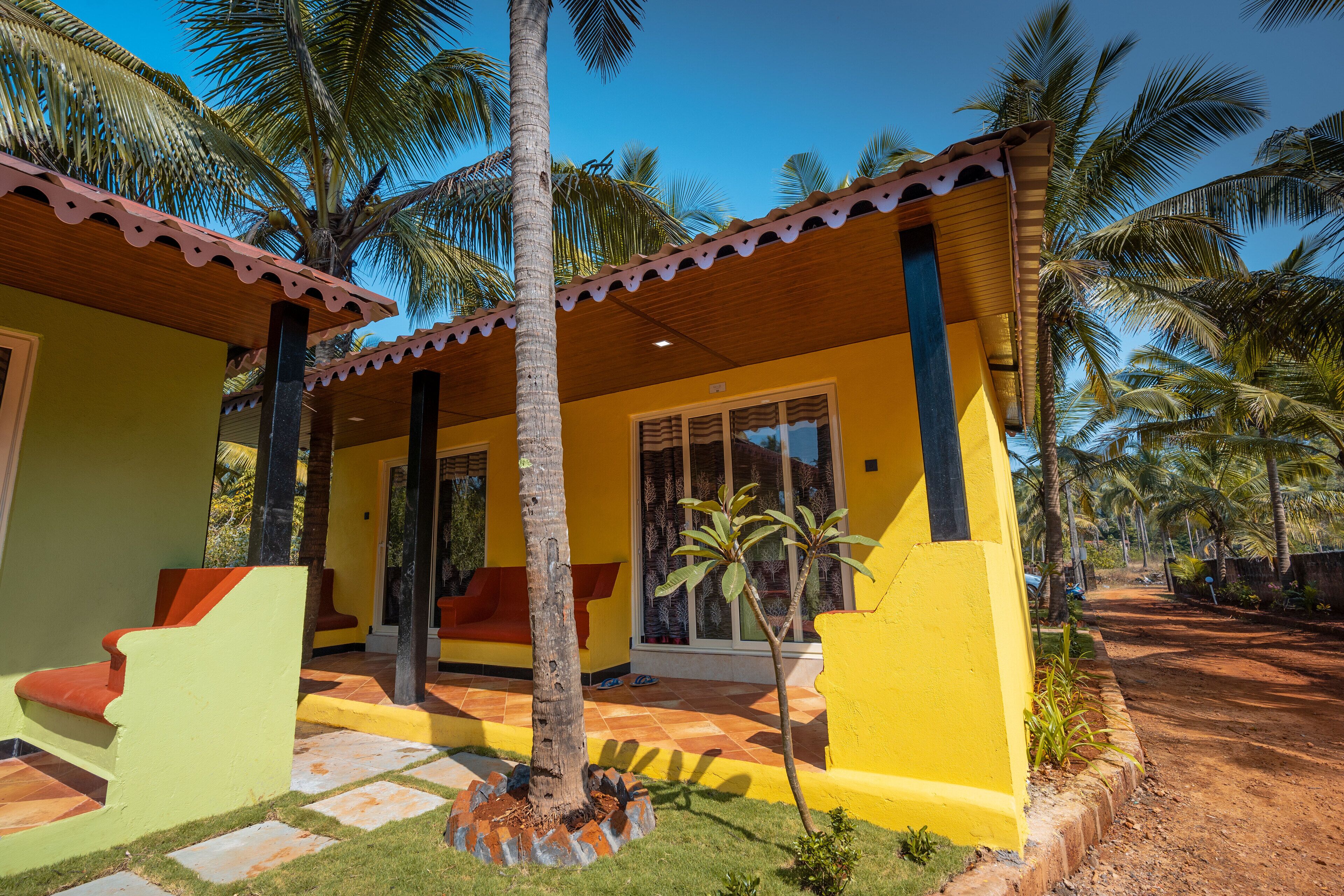 Un edificio amarillo con patio de baldosas, palmeras y mesas al aire libre.