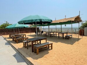 Outdoor seating with tables and benches, umbrellas, and a sandy area. - Kay's By The Sea (Vengurla)