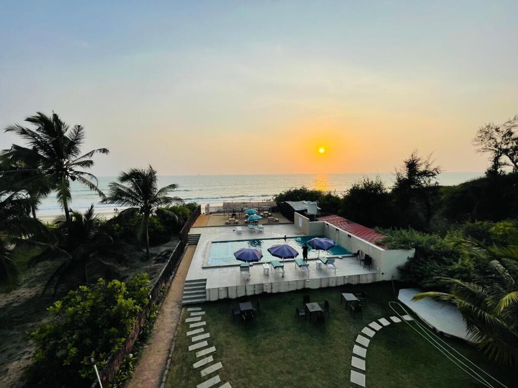 A pool area with lounge chairs and umbrellas, surrounded by palm trees and a view of the ocean at sunset.