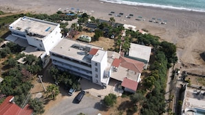 An aerial view of a coastal hotel with multiple buildings, a parking area, and a beach with umbrellas. - Mavi Cennet Camping Pansiyon (Manavgat)